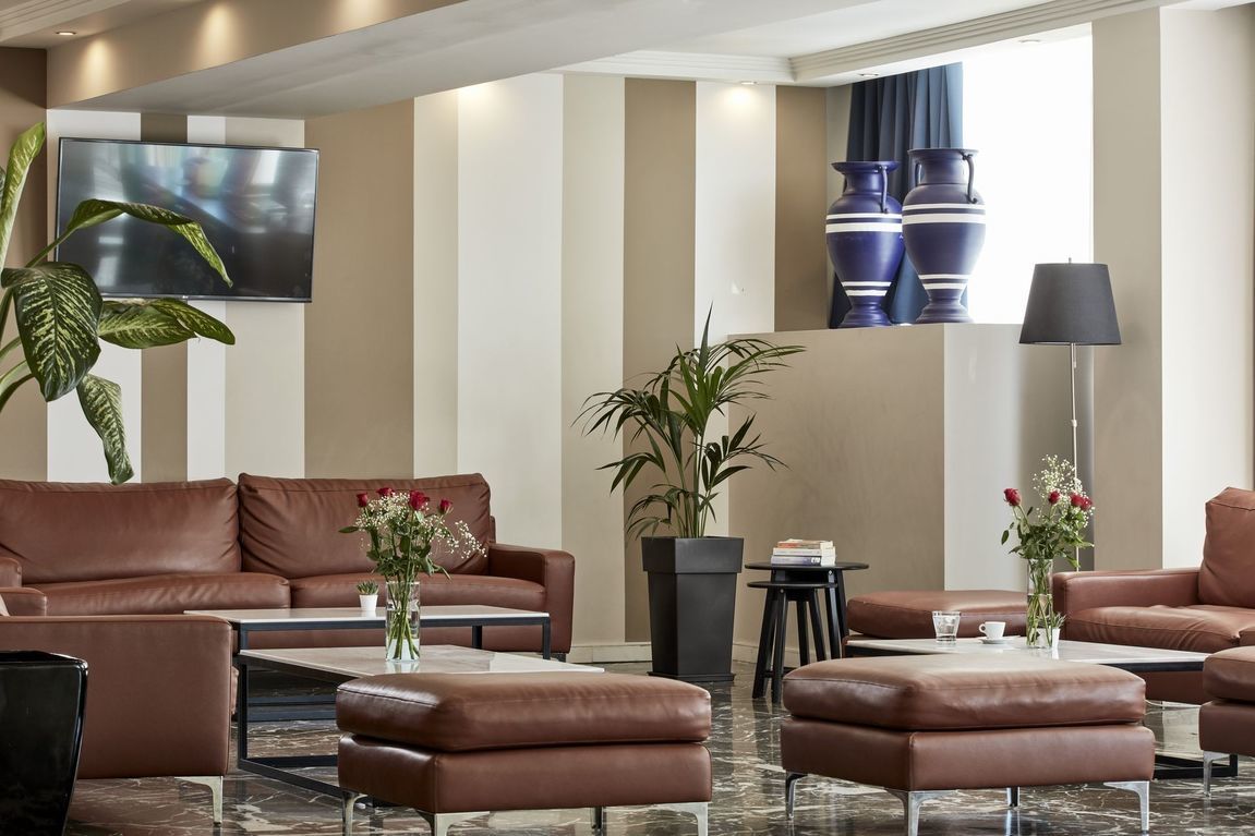 The lounge area of Candia Hotel's lobby featuring tan leather sofas, armchairs and footstools, coffee tables, TV and potted plants